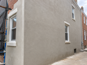 Freshly applied uniform sand-finish stucco coat on the side wall of a Philadelphia rowhome ending at a clean corner bead and window trim
